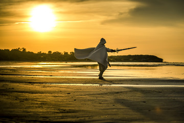 Portrait of assassin in white costume with the sword at the sea. He is posing at the sea during beautiful sunset. Yellow, soft light. - 212038771