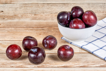 Red plums in a white dish next to a doily and a few plums in front, on a wooden background