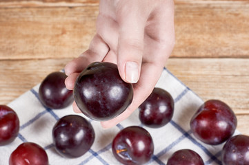female hand holds red plum next to a doily and a few plums behind, on a wooden background