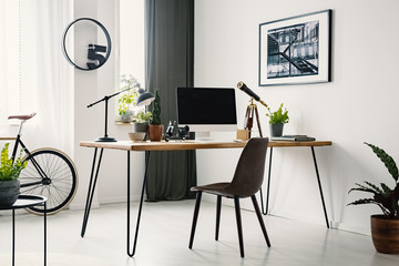 Hairpin desk with mockup monitor, plants and telescope standing in bright room interior with poster...