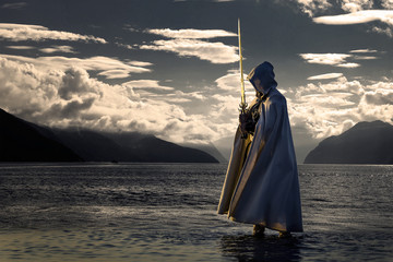 Portrait of assassin in white costume with the sword at the sea. He is posing near water during sunset, soft light. - 212039900
