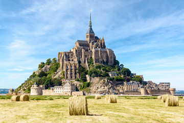 Obraz premium View of the Mont Saint-Michel tidal island, situated in France on the border between Normandy and Brittany, with round bales of straw in a field in the foreground under a blue sky with fibrous clouds.