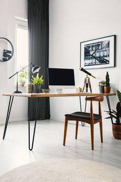Wooden chair standing by the hairpin desk with empty mockup monitor, lamp and fresh potted plants standing in bright living room interior with poster on the wall and window with drape