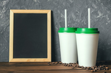 two white plastic coffee cups with a green lid and an inscription frame on a wooden table with scattered coffee beans, front view with copy space - 212041105