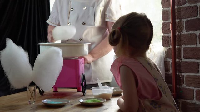 A young male cook makes cotton candy on a special machine, a little girl watches the process and waits for a dessert in a room with brick walls.
