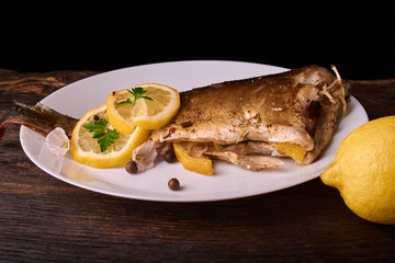 Baked fish with lemon, spices and herbs on white plate on wooden background without head