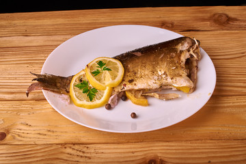 Baked fish with lemon, spices and herbs on white plate on wooden background without head