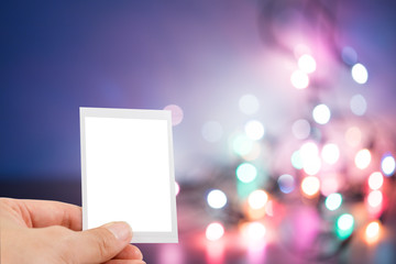 Obraz premium Hand holding instant photo or picture frame with blurred christmas lights background with copy space warm retro vintage tone.