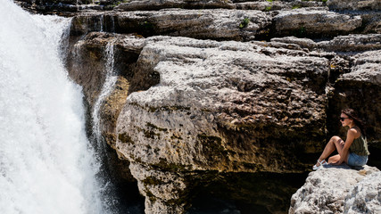 Young girl sitting on the edge of a cliff and looking at the waterfall.