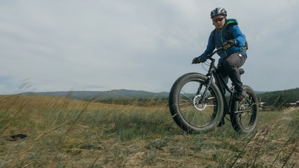 Naklejka na ściany i meble Fat bike also called fatbike or fat-tire bike in summer driving through the hills. The guy is riding a bike along the sand and grass high in the mountains. He performs some tricks and runs dangerously