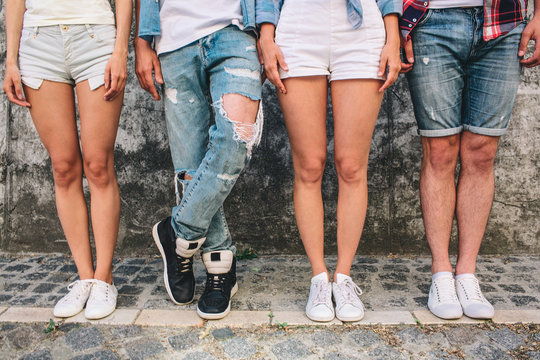 A picture of teens' legs. Three of them are standing straight while man in jeans is holding left leg over right one. Girls wear shorts and one boy as well. They are standing on grey background.