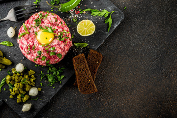 French cuisine, beef steak tartare with raw quail egg yolk, pickled cucumber and onions, fresh herbs, bread, dark rusty background copy space above