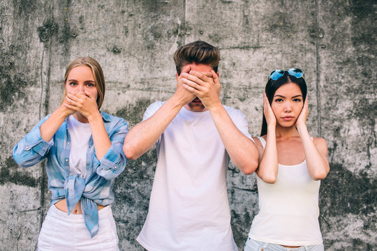 A picture of three young people standing together on grey background. Blonde girl is covering up her mouth with hands. Guy is covering up his eyes. Girl is covering up her ears with hands. They look