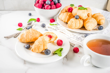 Sweet summer dessert, homemade baked mini croissants with berry jam, served with tea, fresh raspberries, blueberries and mint. On a white marble table, copy space
