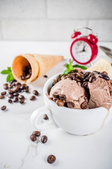 Time for coffee concept, with an alarm clock for hours in the frame. Homemade coffee ice cream, served with coffee beans and mint leaves, with ice cream cones and spoons. White marble background,