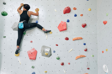 Back view of woman hanging on artificial boulders of climbing wall in gym ascending slowly