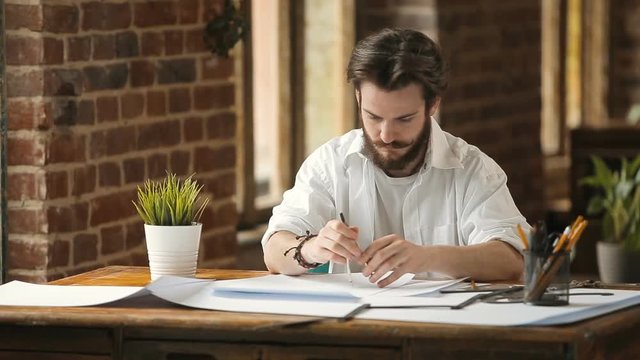Bearded designer dressed in white shirt working with compass, drawings on papers in stylish loft studio