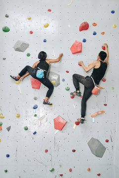 Back view from below of sportive woman and man with bags of talk climbing up artificial boulders on wall in gym
