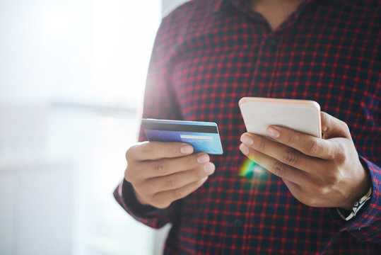 Close-up shot of faceless man in shirt doing purchase online via smartphone with help of credit card