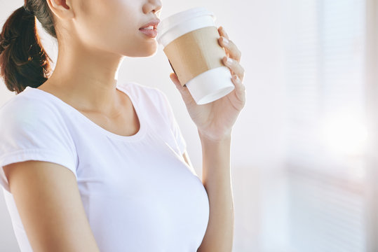 Side view of crop female in white t-shirt holding cup of coffee to go in hand on light blur background