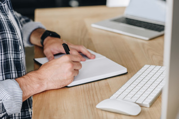 cropped shot of man taking notes in notepad while using desktop computer at workplace