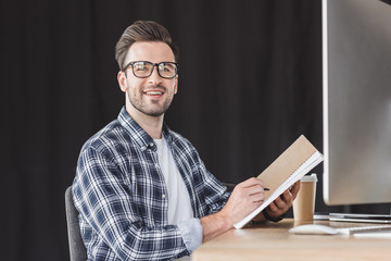 handsome young man in eyeglasses taking notes in notebook and smiling at camera while sitting at workplace
