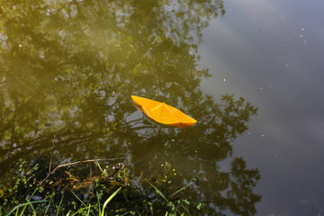 The roots and branches of trees with green grass over the water in the forest. Handmade yellow paper boat in small natural river in the nature. Summer sunny day. Freedom travel creative photo.