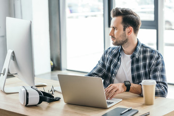 handsome young programmer using laptop and desktop computer at workplace