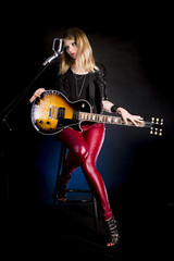 Enigmatic, stylish blonde girl guitarist sits with electric guitar and retro microphone on black background. Woman musician singer dreams, thinks, smiles, shy