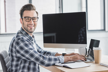 handsome young programmer in eyeglasses smiling at camera while working with laptop and desktop computer