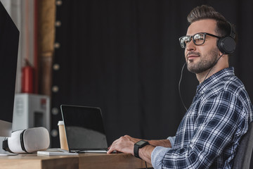 smiling young man in eyeglasses and headphones working with laptop