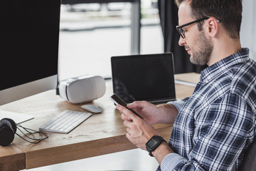 handsome young programmer in eyeglasses using smartphone at workplace
