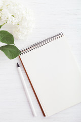 A paper notebook, pencil and flower on white wooden table. Working place and planning concept, top view.