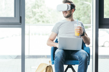 smiling young man holding plastic cup with fresh juice and using virtual reality headset with laptop