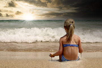 Kind sitzt wartend am Sandstrand und bewundert den Sonnenuntergang