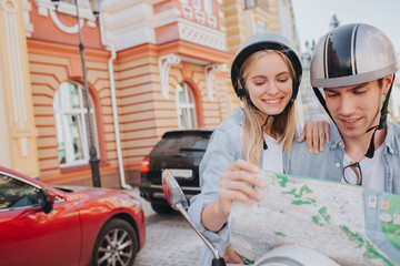 A picture of two travellers sitting on motorcyle and looking on map. Guy is holding it. Girl is smiling and leaning to guy's soulder.