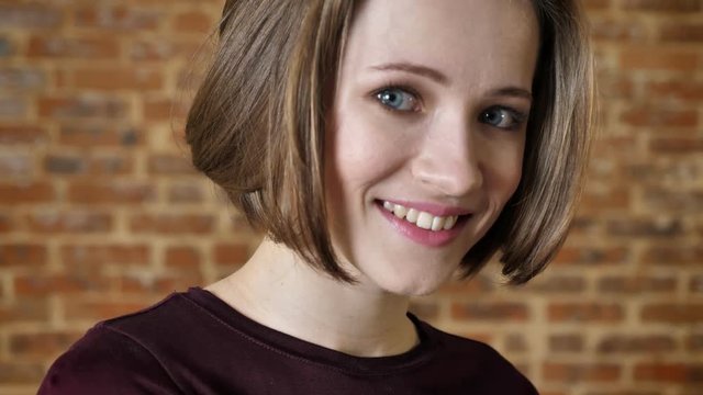 Young attractive shy girl is smiling, look away, watching at camera, brick background