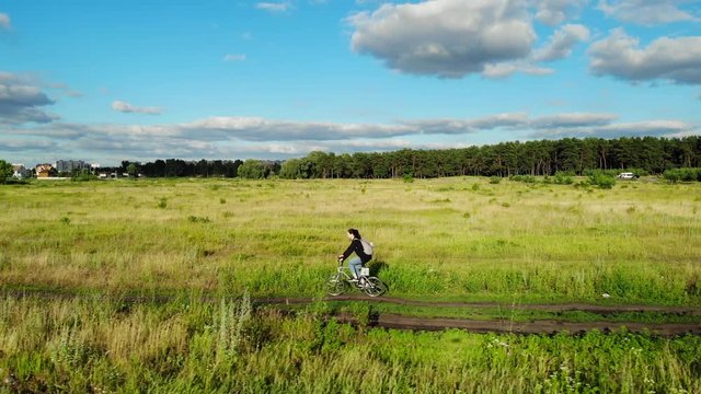 Aerial shot of a girl riding electric bike outdoors on country road in meadow on warm summer day with a drone filming a profile