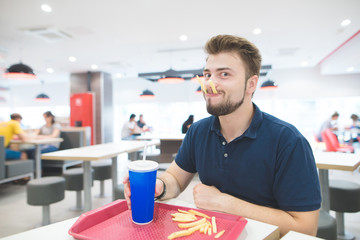Funny man sits in a fast food restaurant with a glass of beverage in his hands and fries in his mouth and looks at the camera. Funny man with a beard eats fast food. Fast Food Concept.