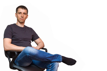 Positive young caucasian man, dressed in a gray shirt, blue jeans and black socks without shoes, sitting on a chair putting his foot on his leg, isolated on white 