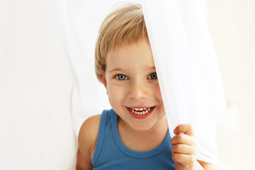 Cute little blond boy sitting on window sill, playing hide-and-seek behind curtain. Portrait of happy adorable three year old child having fun, smiling and laughing. Background, close up, copy space.