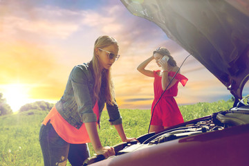 road trip, transport, travel and people concept - young women with open hood of broken car at countryside calling on smartphone to evacuation service over evening sky background