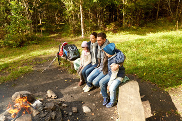 camping, travel, tourism, hike and people concept - happy family sitting on bench at camp fire in woods