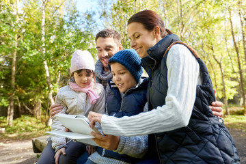 tourism, family and technology concept - happy mother, father, son and daughter sitting on bench with tablet pc computer at camp in woods