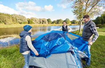 camping, tourism and family concept - happy mother, father and son setting up tent at campsite