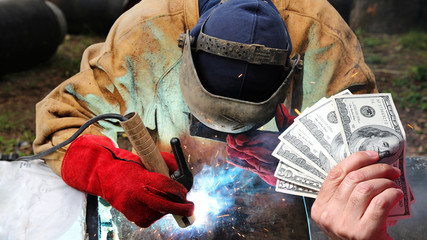 Pipeline Welder Salaries As A Concept / Hand holding US dollar bills in front of an pipeline welder at work with protective equipment - obrazy, fototapety, plakaty