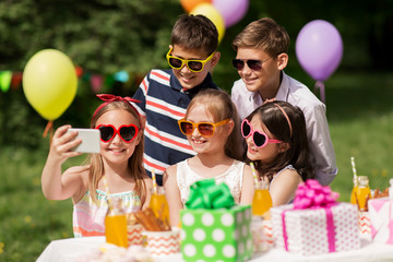 holidays, childhood and technology concept - happy kids in sunglasses taking selfie by smartphone on birthday party at summer garden
