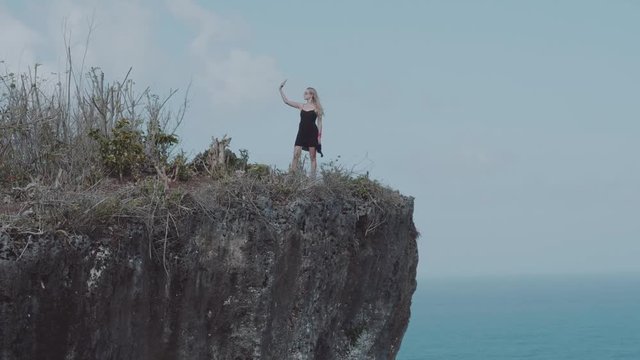 Pretty girl doing selfie photo while standing on the cliff with amazing ocean and cloudy sky view - video in slow motion