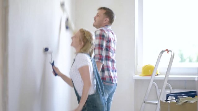 Young family makes repairs in new flat. Couple - painting wall in apartment. Man and woman doing overhaul of house. Funny wife and husband having good mood - making decorate room and dancing.