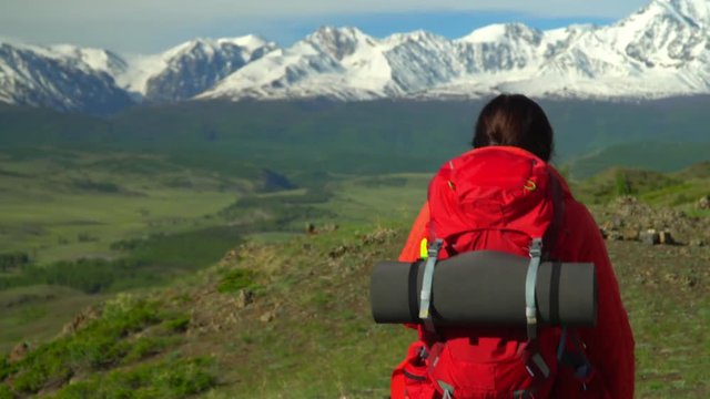 The woman with a red backpack goes on a slope of the mountain and looks at snow mountains.